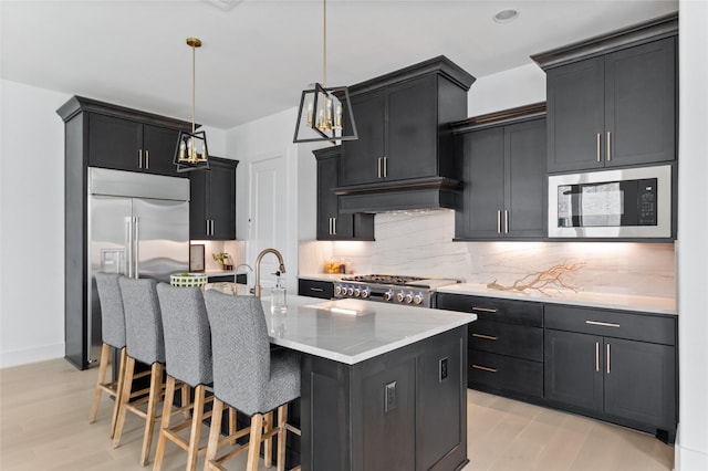 kitchen with a kitchen island with sink, hanging light fixtures, decorative backsplash, appliances with stainless steel finishes, and light wood-type flooring