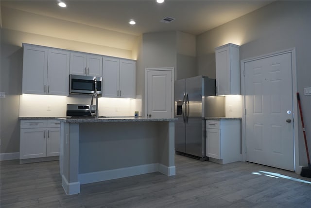 kitchen featuring appliances with stainless steel finishes, light hardwood / wood-style flooring, stone counters, white cabinetry, and an island with sink