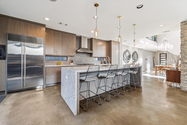 kitchen featuring a kitchen island with sink, built in refrigerator, wall chimney exhaust hood, decorative light fixtures, and tasteful backsplash