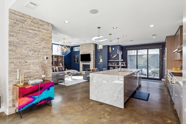 kitchen featuring sink, decorative light fixtures, a fireplace, light stone counters, and an island with sink