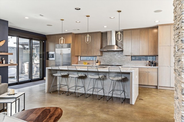 kitchen with hanging light fixtures, an island with sink, built in appliances, and wall chimney exhaust hood