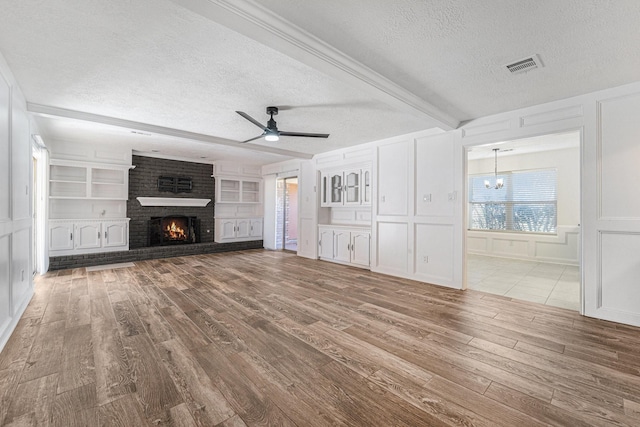 unfurnished living room featuring a brick fireplace, built in shelves, ceiling fan with notable chandelier, a textured ceiling, and light hardwood / wood-style floors