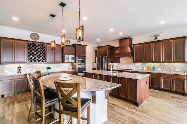 kitchen with light stone countertops, stainless steel appliances, decorative light fixtures, a center island with sink, and custom exhaust hood