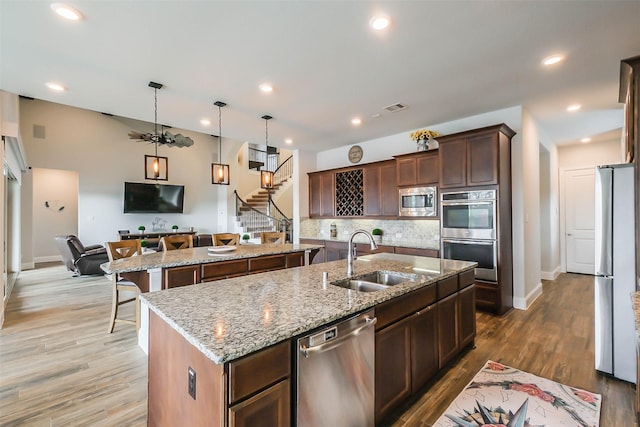 kitchen with a center island with sink, sink, hanging light fixtures, light stone counters, and stainless steel appliances