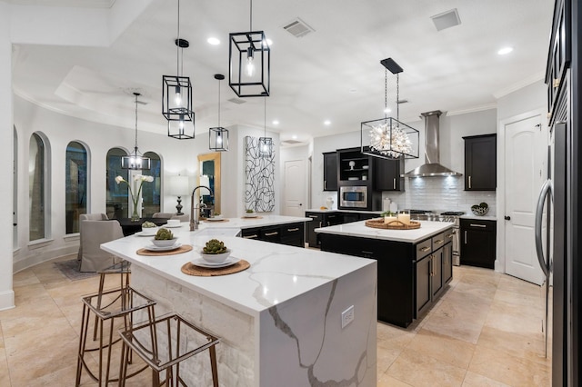 kitchen featuring a kitchen island with sink, stainless steel appliances, wall chimney range hood, pendant lighting, and decorative backsplash