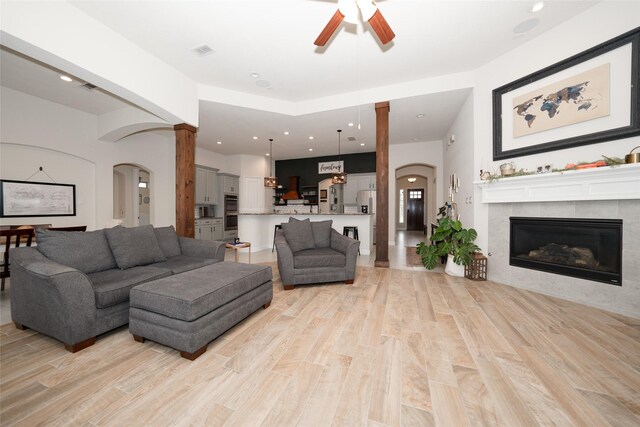 living room with ceiling fan, light wood-type flooring, a tile fireplace, and decorative columns