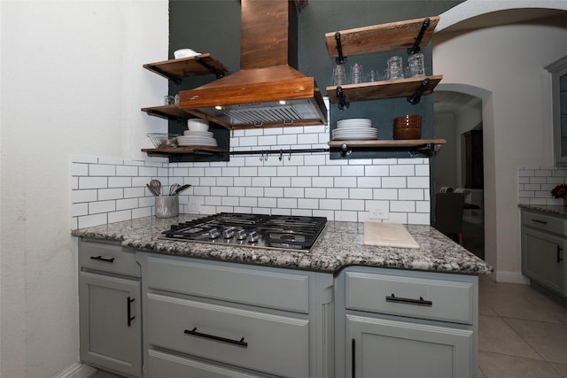 kitchen with stainless steel gas cooktop, backsplash, dark stone countertops, light tile patterned flooring, and custom exhaust hood