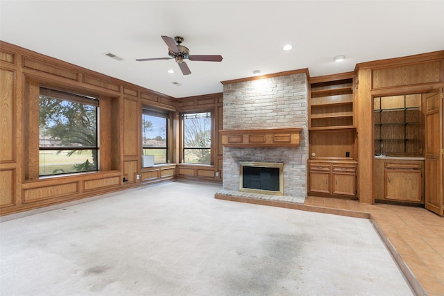 unfurnished living room with wood walls, a brick fireplace, built in features, ceiling fan, and light carpet