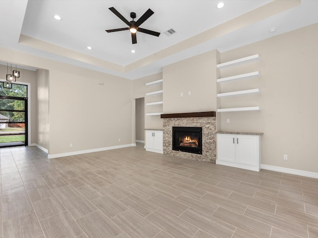 unfurnished living room with ceiling fan, a stone fireplace, and a tray ceiling