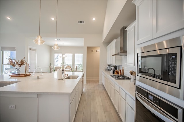 kitchen with sink, stainless steel appliances, hanging light fixtures, wall chimney range hood, and an island with sink