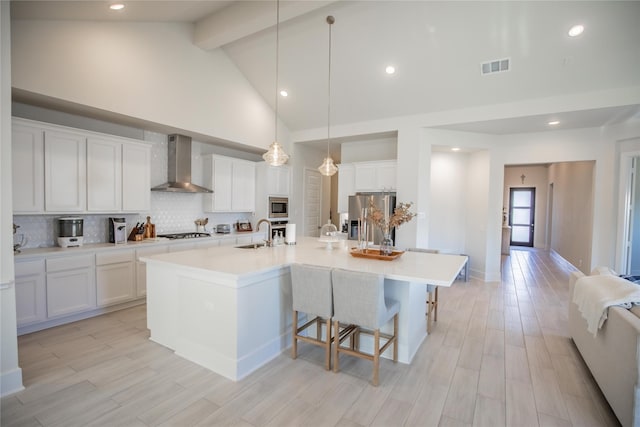 kitchen with beam ceiling, wall chimney exhaust hood, decorative light fixtures, a center island with sink, and white cabinets