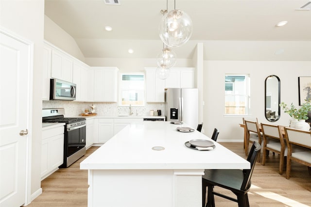 kitchen featuring vaulted ceiling, decorative light fixtures, a kitchen island with sink, white cabinets, and appliances with stainless steel finishes