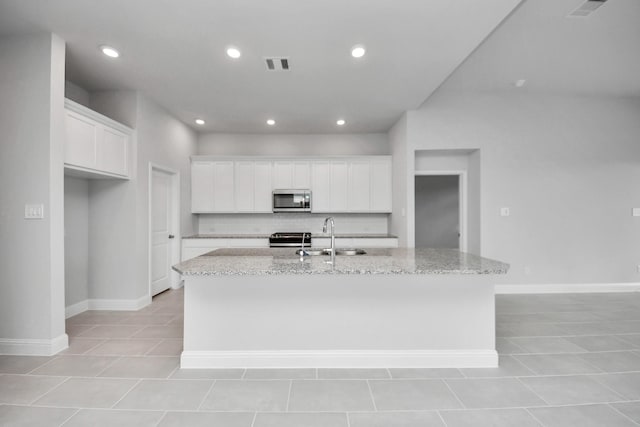 kitchen with light stone counters, stainless steel appliances, a sink, visible vents, and an island with sink