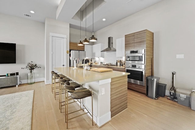 kitchen with appliances with stainless steel finishes, decorative light fixtures, a kitchen breakfast bar, a kitchen island with sink, and wall chimney exhaust hood