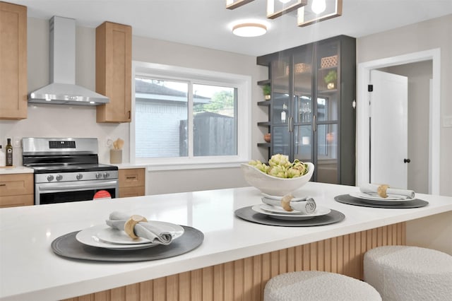 kitchen featuring light brown cabinetry, wall chimney range hood, stainless steel electric stove, and a breakfast bar area