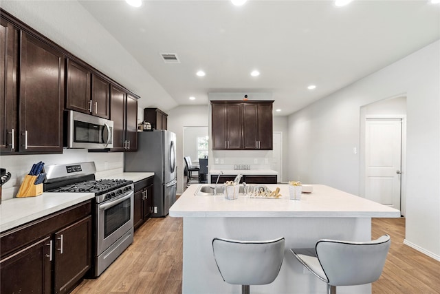 kitchen with stainless steel appliances, an island with sink, a kitchen bar, dark brown cabinets, and light wood-type flooring