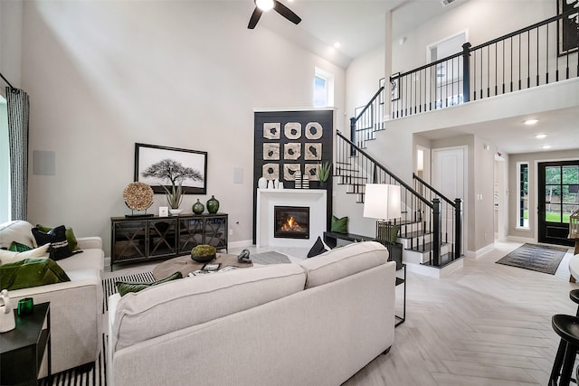 living room with a high ceiling, a ceiling fan, a glass covered fireplace, baseboards, and stairs