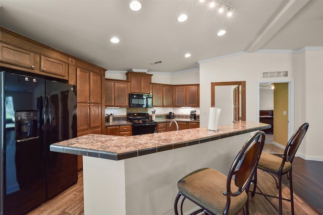 kitchen featuring tile counters, a kitchen breakfast bar, ornamental molding, black appliances, and hardwood / wood-style flooring