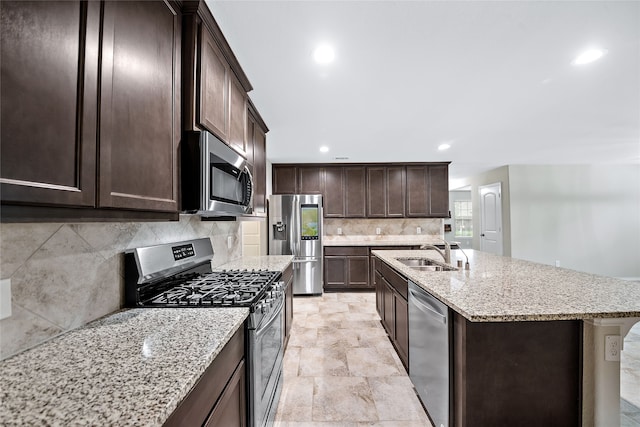 kitchen with light stone countertops, stainless steel appliances, a kitchen island with sink, dark brown cabinets, and sink