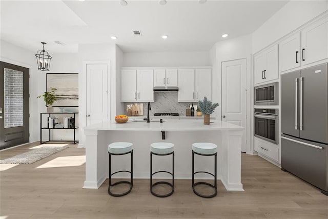 kitchen with stainless steel appliances, backsplash, an island with sink, white cabinets, and light wood-type flooring