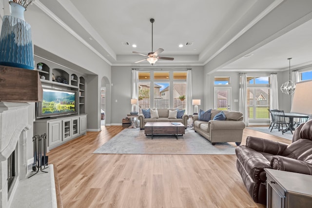 living room featuring built in shelves, ceiling fan with notable chandelier, a raised ceiling, crown molding, and light hardwood / wood-style flooring