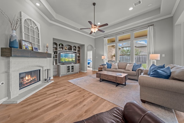 living room with light wood-type flooring, ornamental molding, built in shelves, a tray ceiling, and ceiling fan