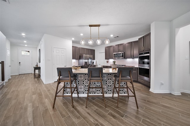 kitchen featuring dark brown cabinetry, pendant lighting, stainless steel appliances, a kitchen island with sink, and decorative backsplash