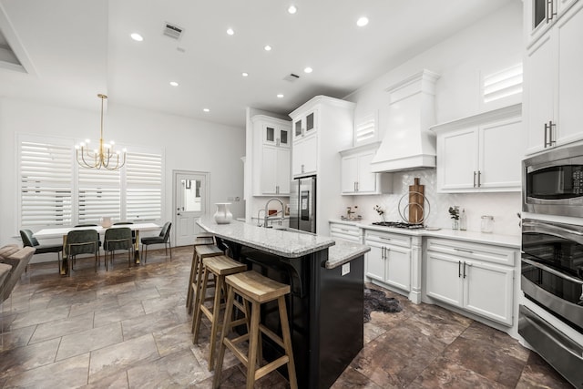 kitchen featuring premium range hood, white cabinets, an island with sink, decorative light fixtures, and stainless steel appliances