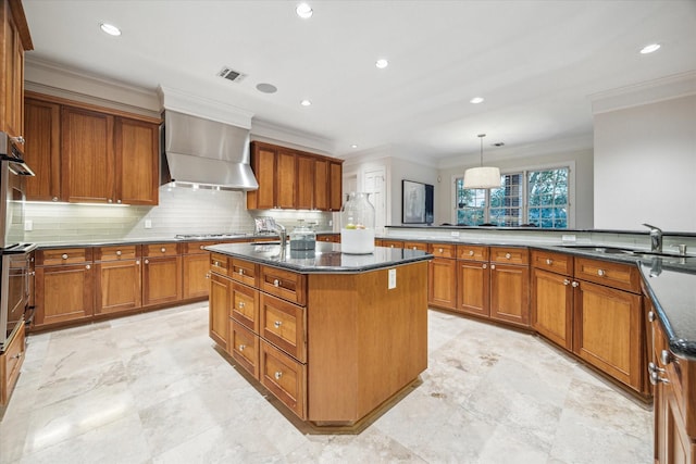 kitchen featuring pendant lighting, wall chimney exhaust hood, dark stone countertops, a center island with sink, and crown molding