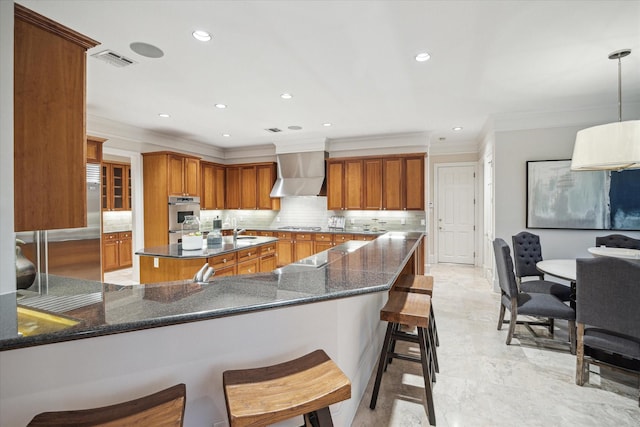 kitchen featuring wall chimney range hood, dark stone counters, hanging light fixtures, kitchen peninsula, and a breakfast bar area