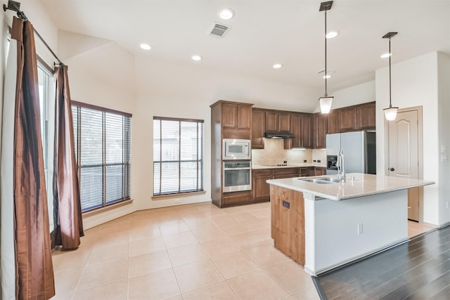 kitchen with sink, appliances with stainless steel finishes, hanging light fixtures, light stone counters, and an island with sink