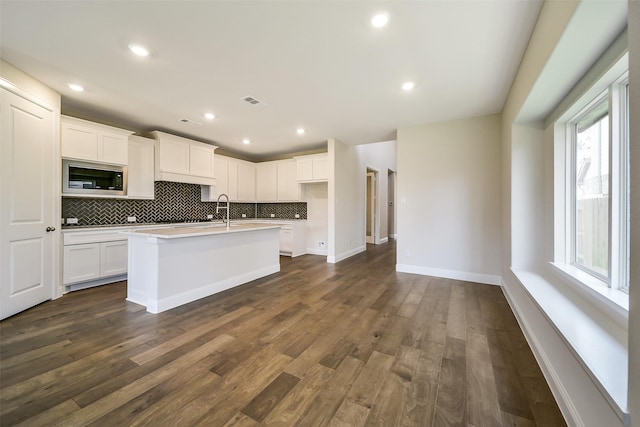 kitchen with stainless steel microwave, backsplash, an island with sink, white cabinets, and light countertops