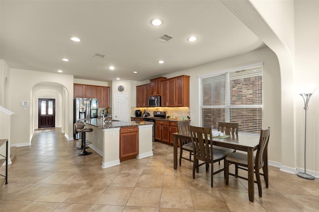 kitchen with stainless steel appliances, light tile patterned floors, a center island with sink, dark stone countertops, and a breakfast bar area