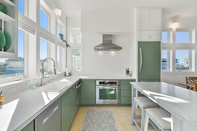 kitchen with green cabinets, light countertops, stainless steel appliances, wall chimney exhaust hood, and a sink