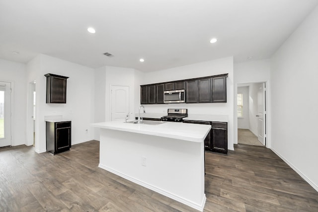 kitchen with sink, dark wood-type flooring, stainless steel appliances, dark brown cabinets, and a center island with sink