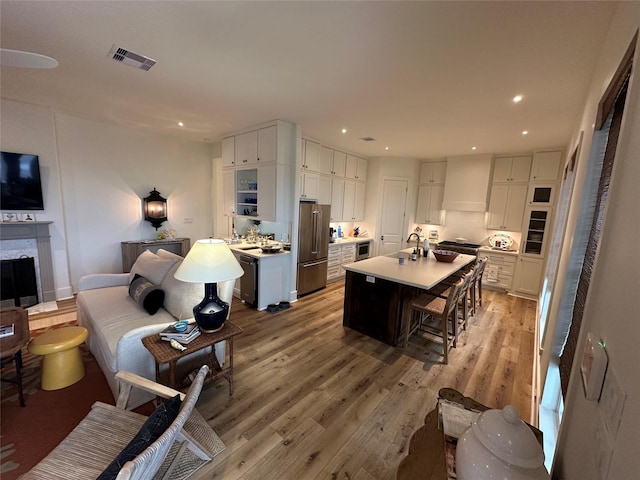 kitchen with high end refrigerator, light wood-type flooring, premium range hood, a kitchen island with sink, and white cabinetry