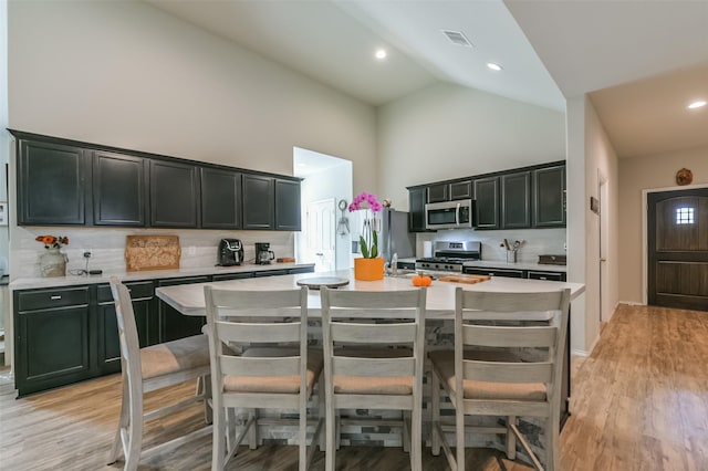 kitchen with high vaulted ceiling, a kitchen breakfast bar, an island with sink, appliances with stainless steel finishes, and tasteful backsplash