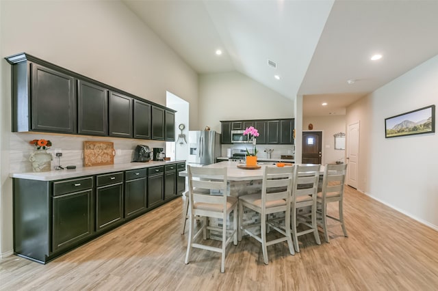 kitchen with a kitchen island with sink, high vaulted ceiling, light wood-type flooring, appliances with stainless steel finishes, and a kitchen bar