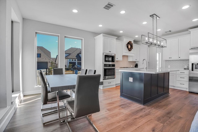 kitchen with stainless steel appliances, white cabinetry, hardwood / wood-style floors, an island with sink, and hanging light fixtures