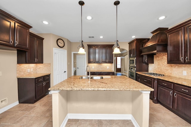 kitchen featuring premium range hood, a kitchen island with sink, sink, decorative light fixtures, and dark brown cabinets