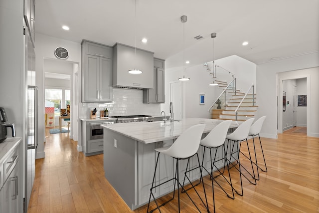 kitchen with light stone countertops, decorative light fixtures, light wood-type flooring, a kitchen island with sink, and gray cabinets