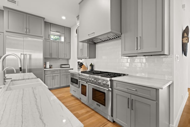 kitchen with light stone counters, high end appliances, wall chimney exhaust hood, light wood-type flooring, and gray cabinets