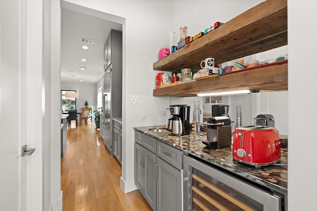 bar featuring built in fridge, dark stone countertops, light hardwood / wood-style floors, beverage cooler, and gray cabinets