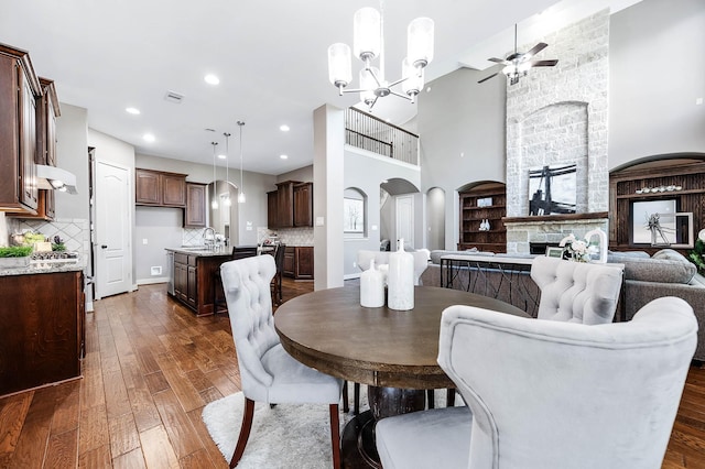 dining room with a stone fireplace, dark hardwood / wood-style floors, built in shelves, ceiling fan with notable chandelier, and sink