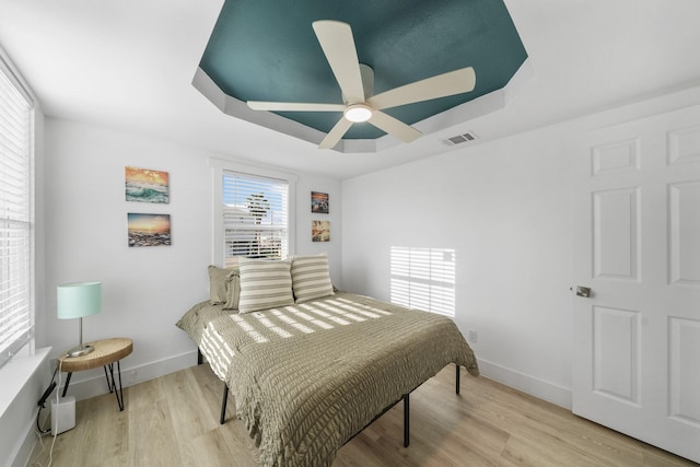 bedroom featuring a tray ceiling, multiple windows, ceiling fan, and light hardwood / wood-style flooring