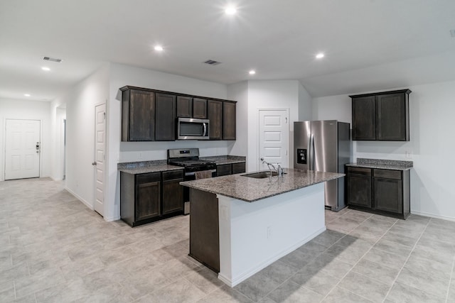 kitchen featuring appliances with stainless steel finishes, an island with sink, dark stone counters, dark brown cabinetry, and sink