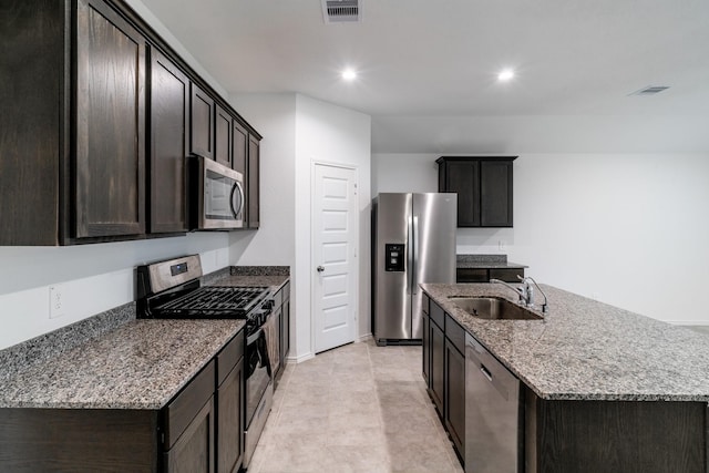 kitchen featuring a center island with sink, appliances with stainless steel finishes, light stone counters, sink, and dark brown cabinets