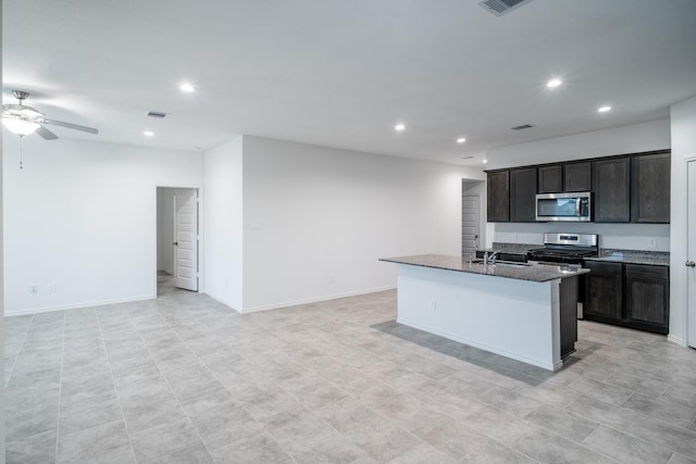 kitchen featuring a kitchen island with sink, dark stone counters, appliances with stainless steel finishes, ceiling fan, and sink