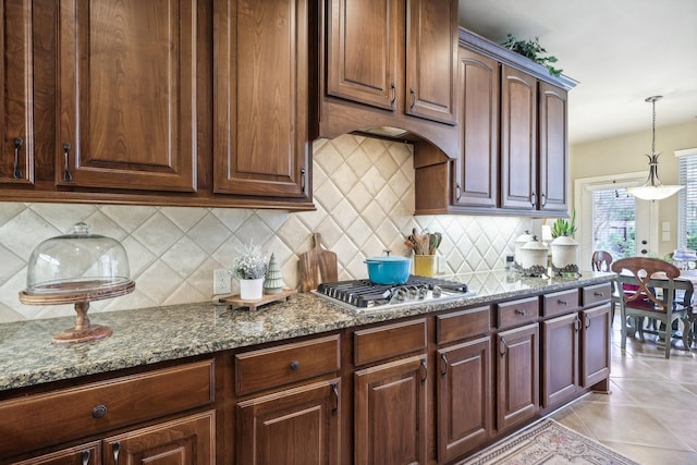 kitchen featuring dark stone countertops, light tile patterned floors, stainless steel gas stovetop, decorative light fixtures, and backsplash