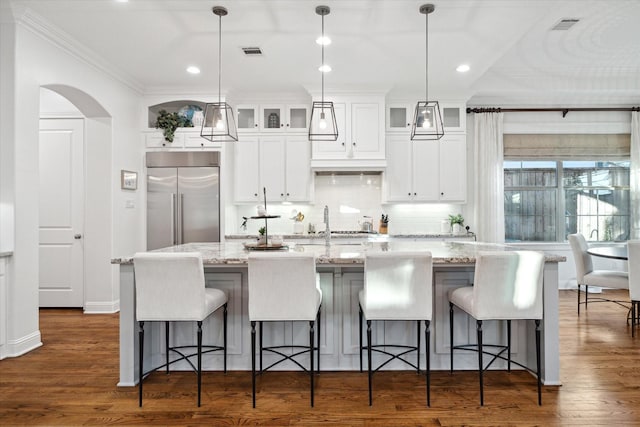 kitchen with light stone counters, dark hardwood / wood-style flooring, stainless steel built in fridge, an island with sink, and pendant lighting